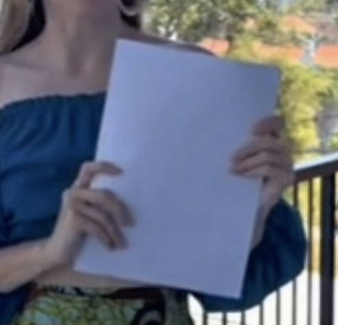 A person in a blue top holding a blank sheet on a balcony, with both hands overlapping its edges.