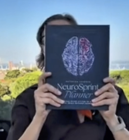 A person outdoors holding a dark blue book with a brain illustration and NeuroSprint Planner text in front of their face.